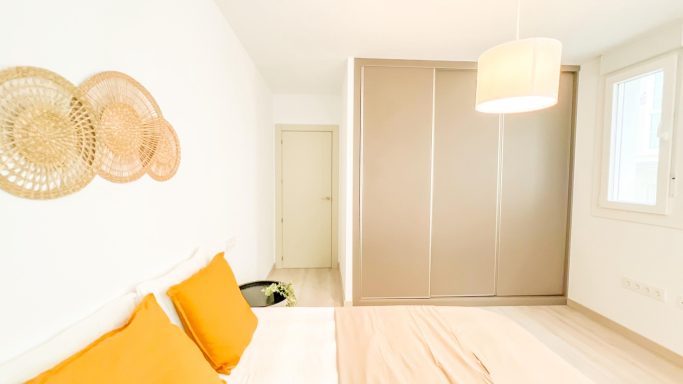 Habitación luminosa con cama blanca y cojines naranjas, armario y decoración sencilla.