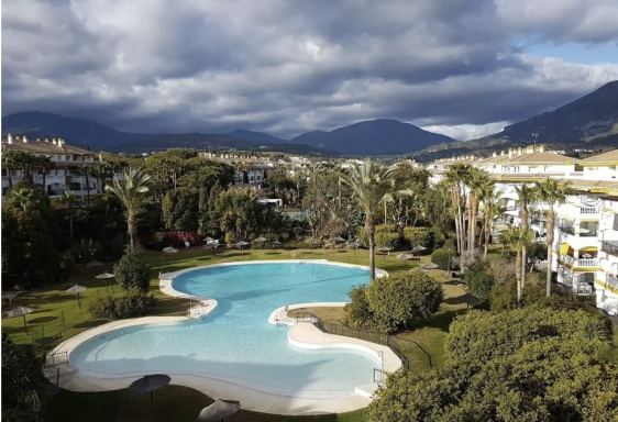 Paisaje de un complejo con piscina y jardines, rodeado de montañas y nubes.