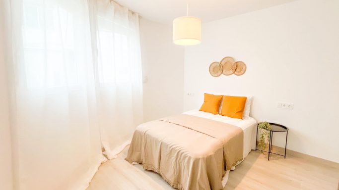 Habitación luminosa con cama, cojines naranjas y decoraciones en la pared.