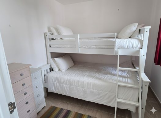 Habitación con literas blancas, cama doble abajo y cama individual arriba, con muebles sencillos.