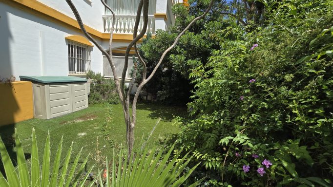 Jardín con vegetación verde, un árbol y un cobertizo en un entorno residencial.